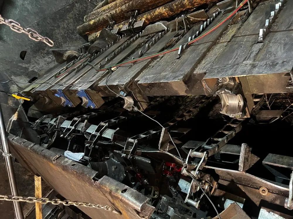 Gros plan sur une lourde machine de convoyage industrielle, composée de plaques métalliques sombres et de chaînes mécaniques, dans un atelier.