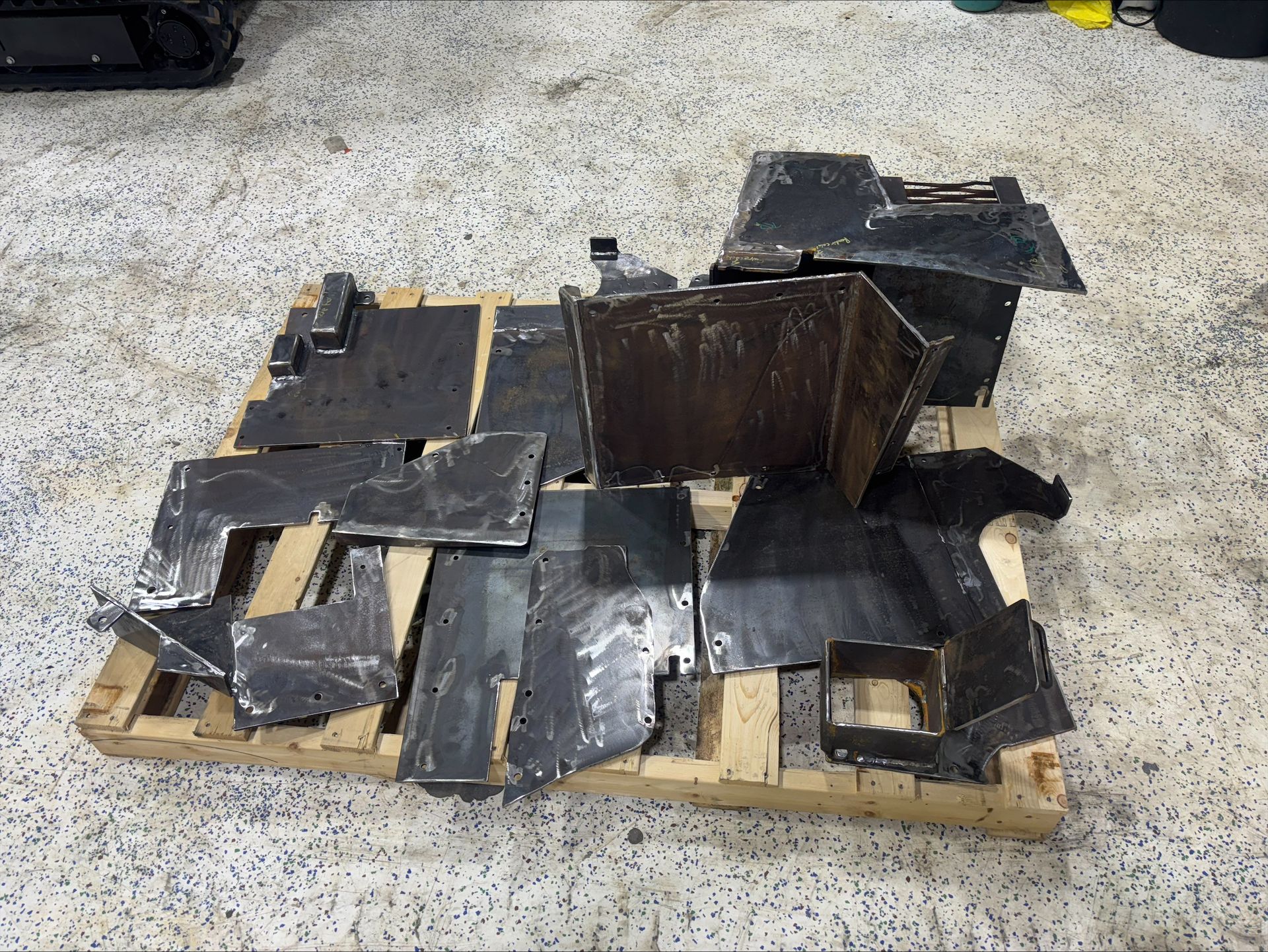 Une collection de plaques de métal sombres et plates et de morceaux découpés, disposés sur une palette d'expédition en bois, elle-même posée sur un sol en béton.