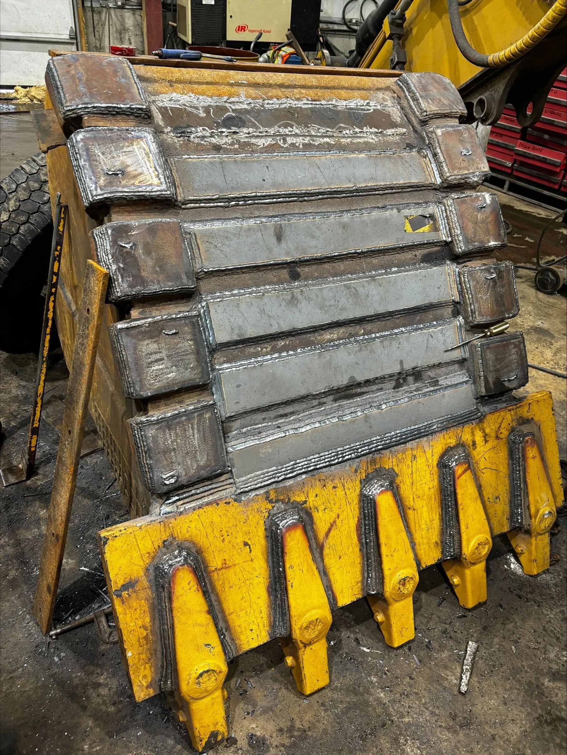 Un godet d'excavatrice jaune, avec des bandes d'usure en acier nouvellement soudées et des dents sur le bord avant, se trouve dans un atelier.