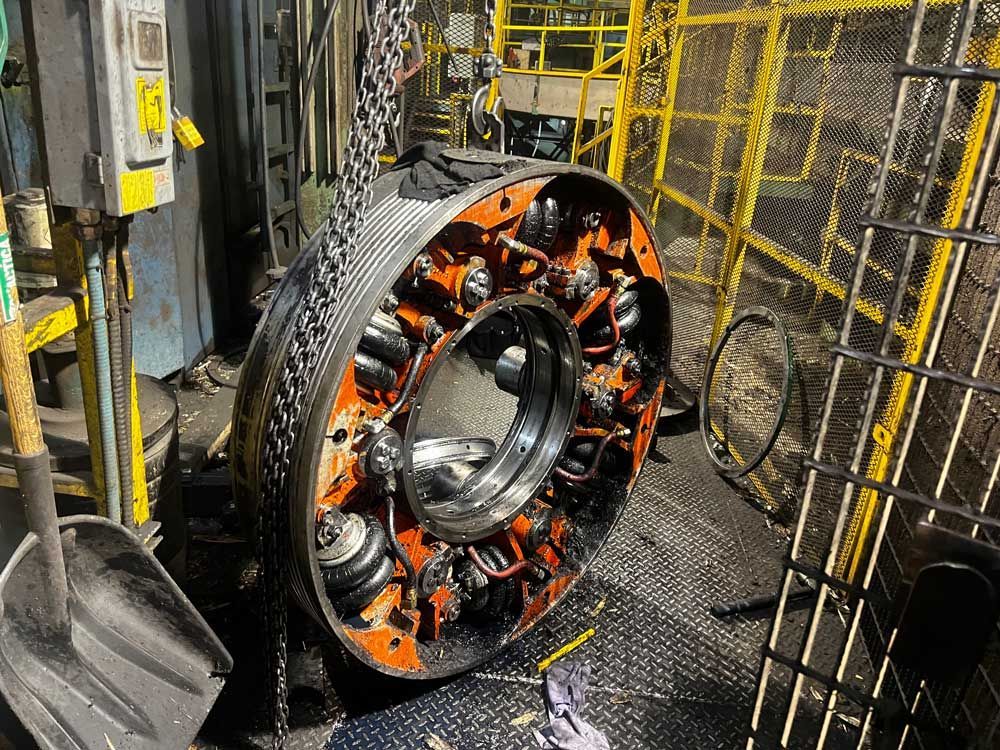 Un grand composant industriel orange et métallique en forme d'anneau est suspendu à un palan à chaîne dans un environnement d'usine.