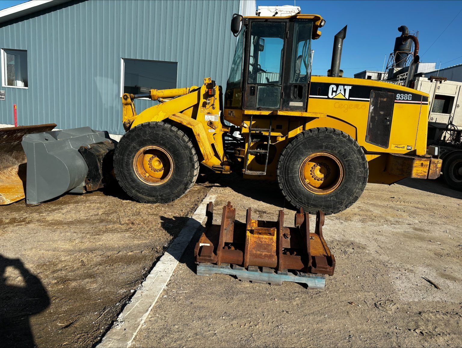 Une chargeuse frontale Caterpillar jaune est garée sur du gravier, avec un accessoire métallique rouillé posé sur une palette au premier plan.