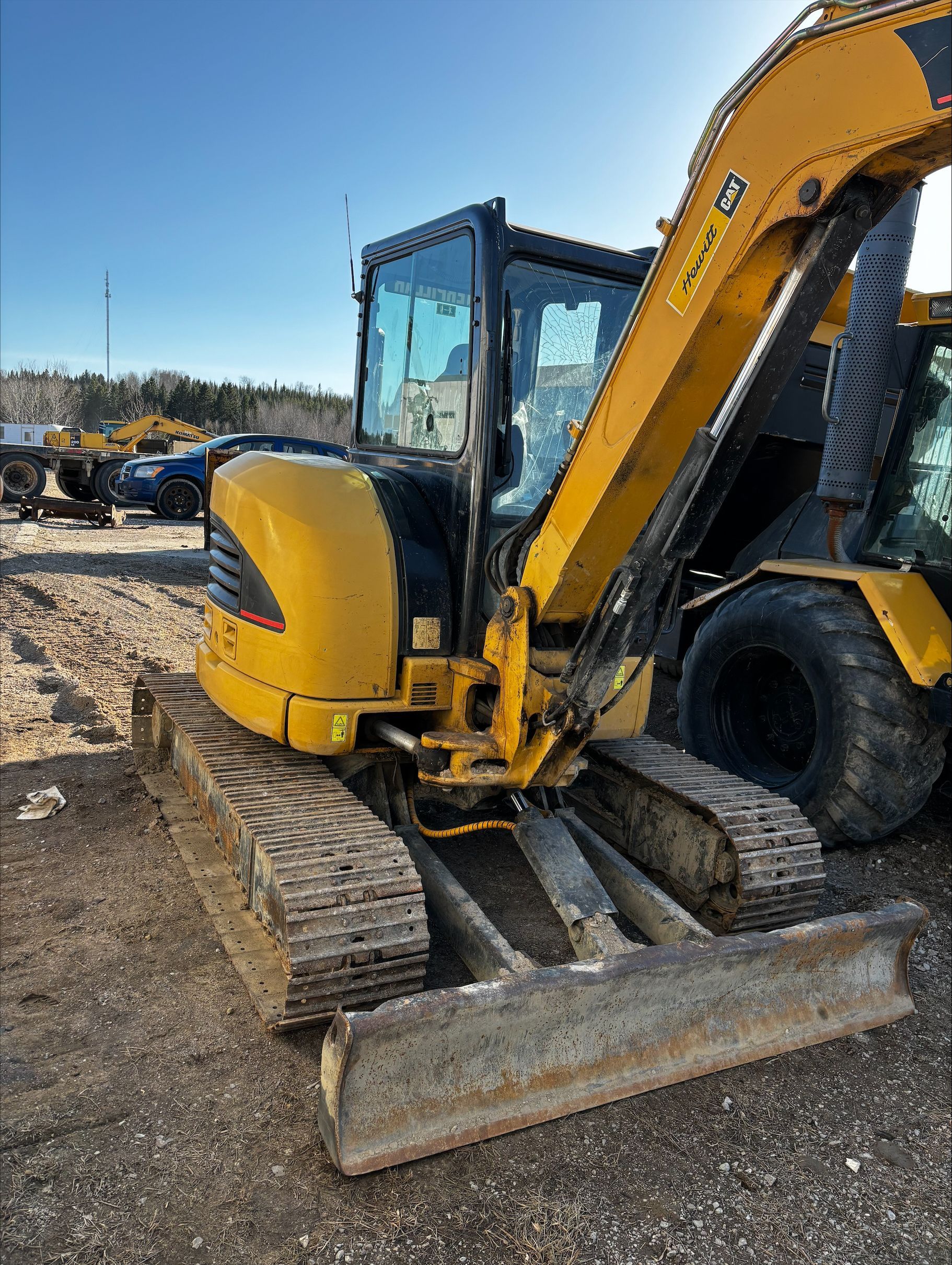 Une mini-pelle Caterpillar jaune est posée sur un terrain gravillonné sous un ciel bleu azur.
