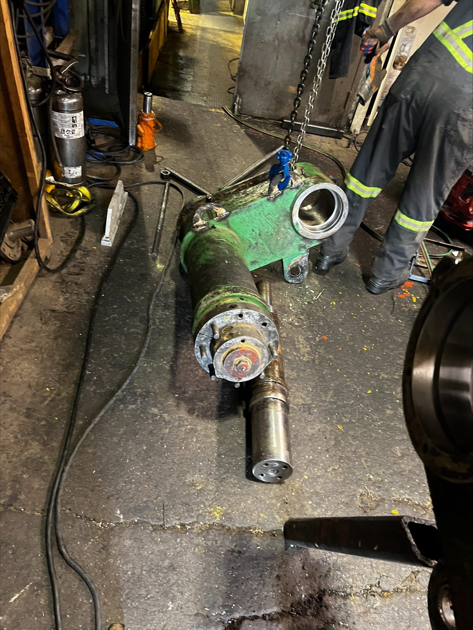 Un ouvrier portant un gilet haute visibilité inspecte un composant industriel hydraulique vert robuste sur le sol d'un atelier.