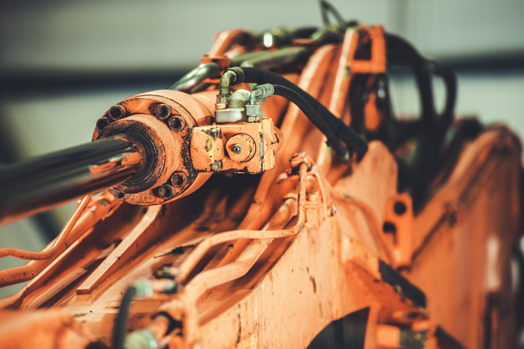 Gros plan sur un vérin hydraulique orange patiné, monté sur un engin de chantier lourd, avec des tubes métalliques fixés.