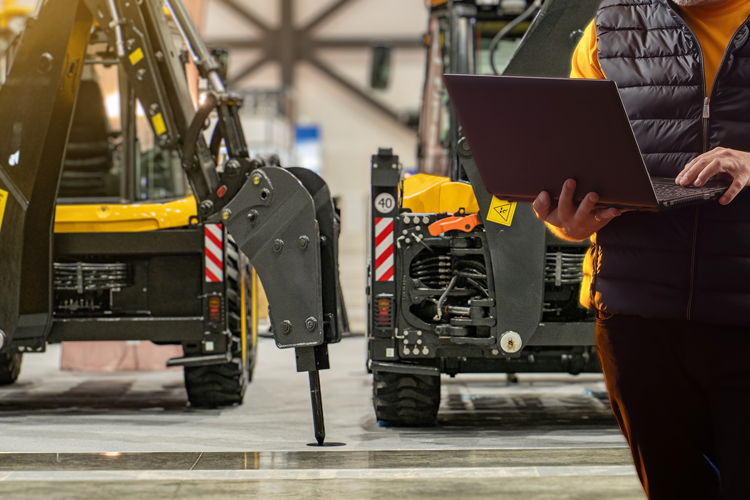 Une personne en gilet se tient debout dans un entrepôt, un ordinateur portable à la main, devant des engins de chantier jaunes.