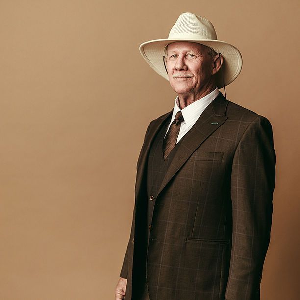 A man in a suit and hat is standing in front of a brown wall