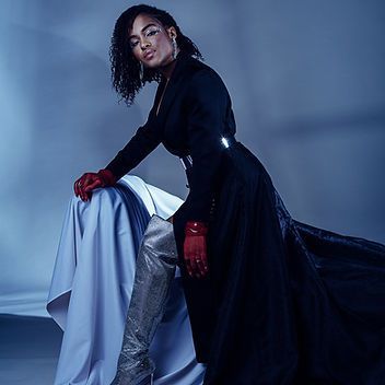 A woman in a black dress and red gloves is sitting on a bed.