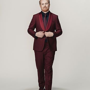 A man in a maroon suit and tie is standing in front of a white background.