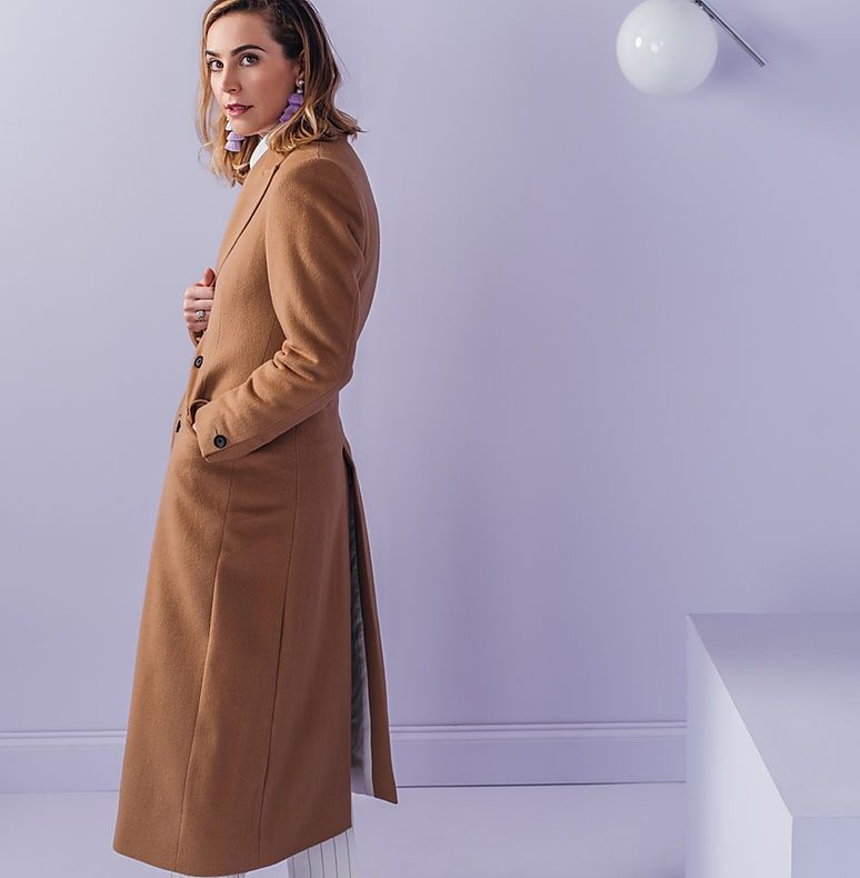 A woman in a long brown coat is standing in front of a purple wall