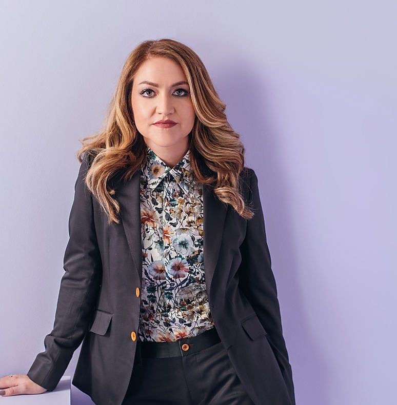 A woman in a suit is leaning against a purple wall