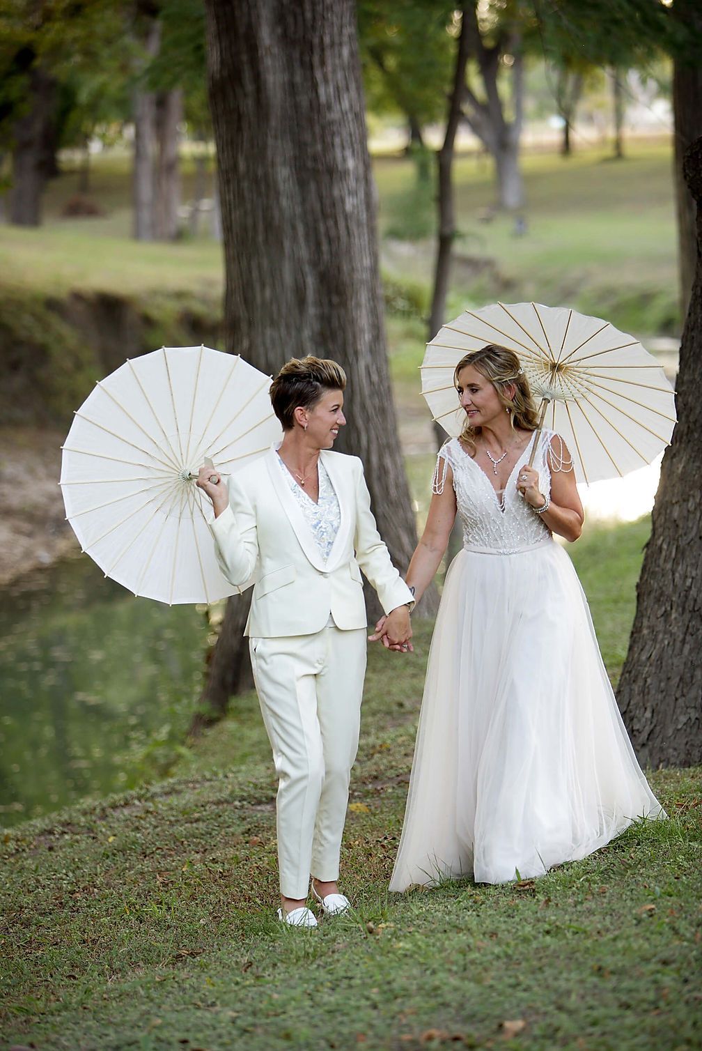 Two women in white dresses are holding white umbrellas in a park.