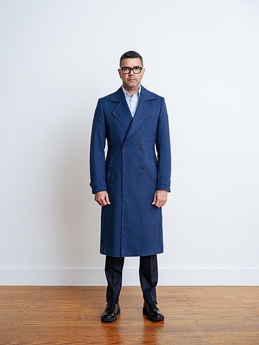 A man wearing a blue coat and glasses is standing on a wooden floor.