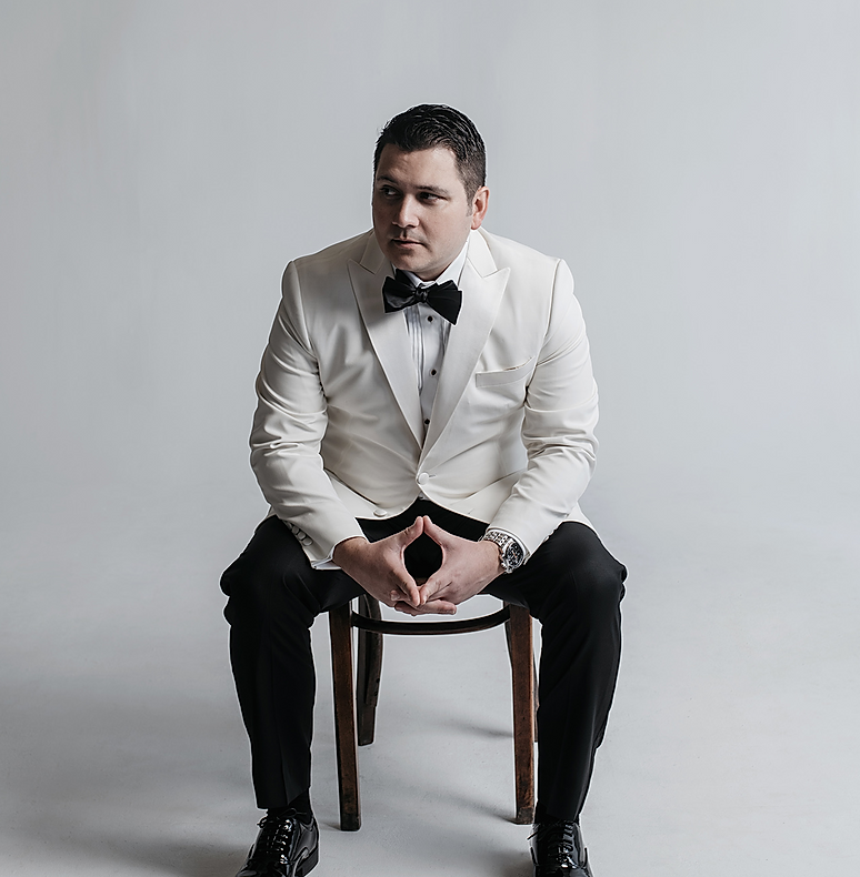 A man in a tuxedo is sitting on a chair with his hands folded