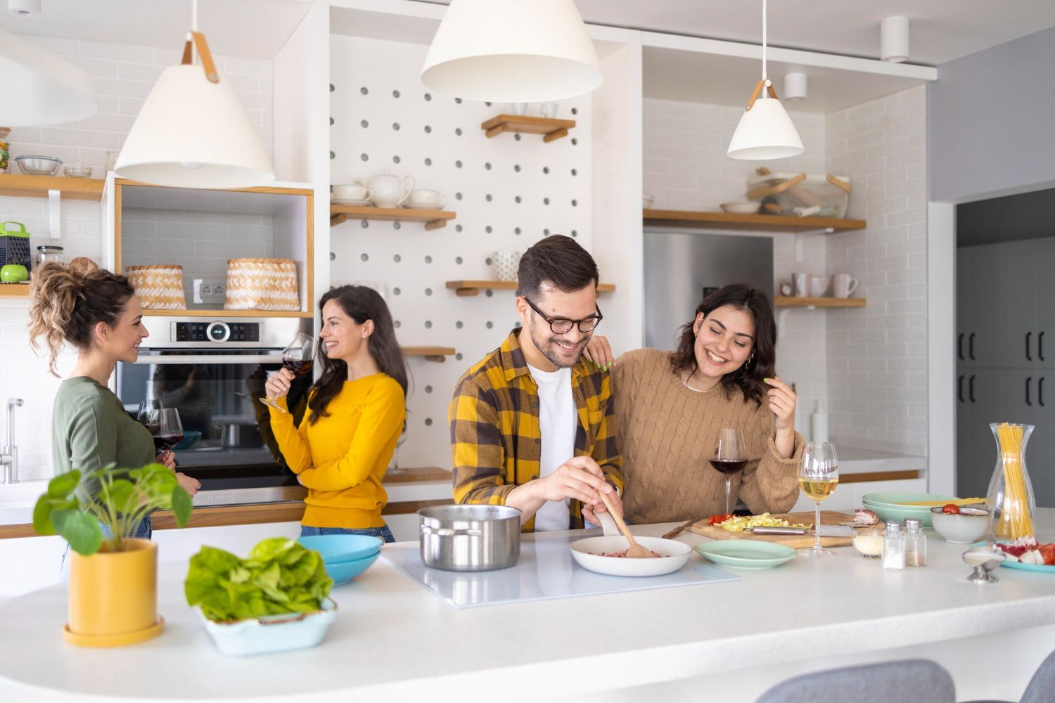 Four friends cooking and enjoying wine in a modern kitchen with white countertops, light wood shelving, and pendant lights.