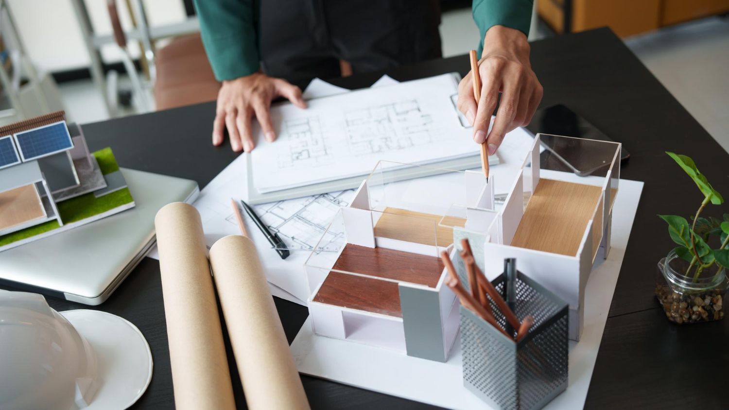 Architect at desk, reviewing blueprints, with model building, color samples, and tools.