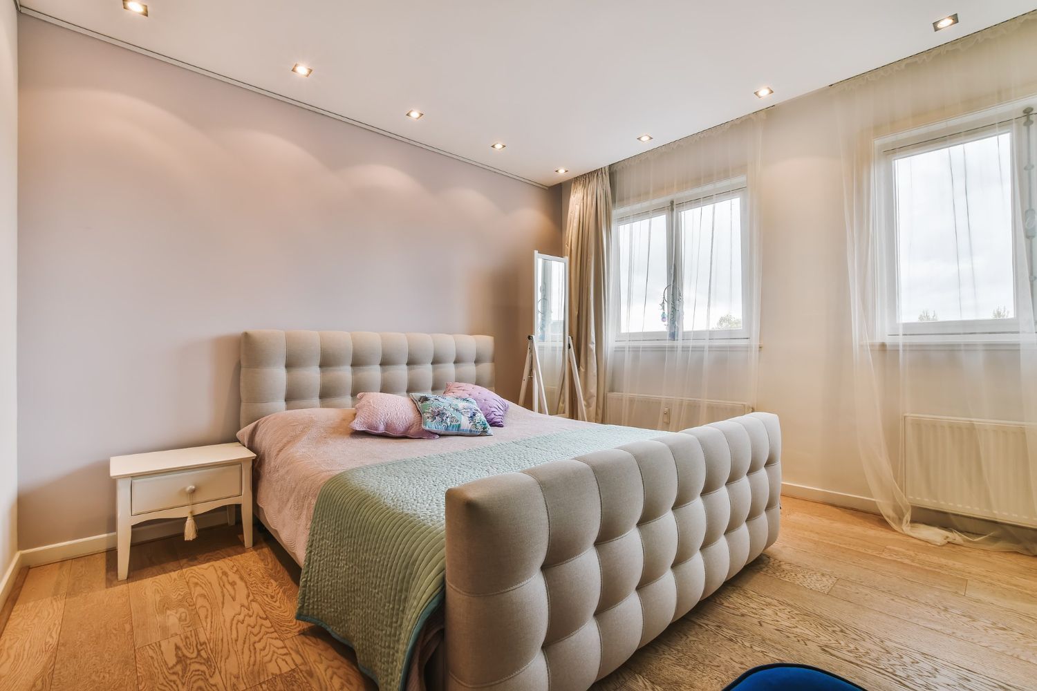 Bedroom with tufted beige bed, nightstand, windows, and wooden floor.