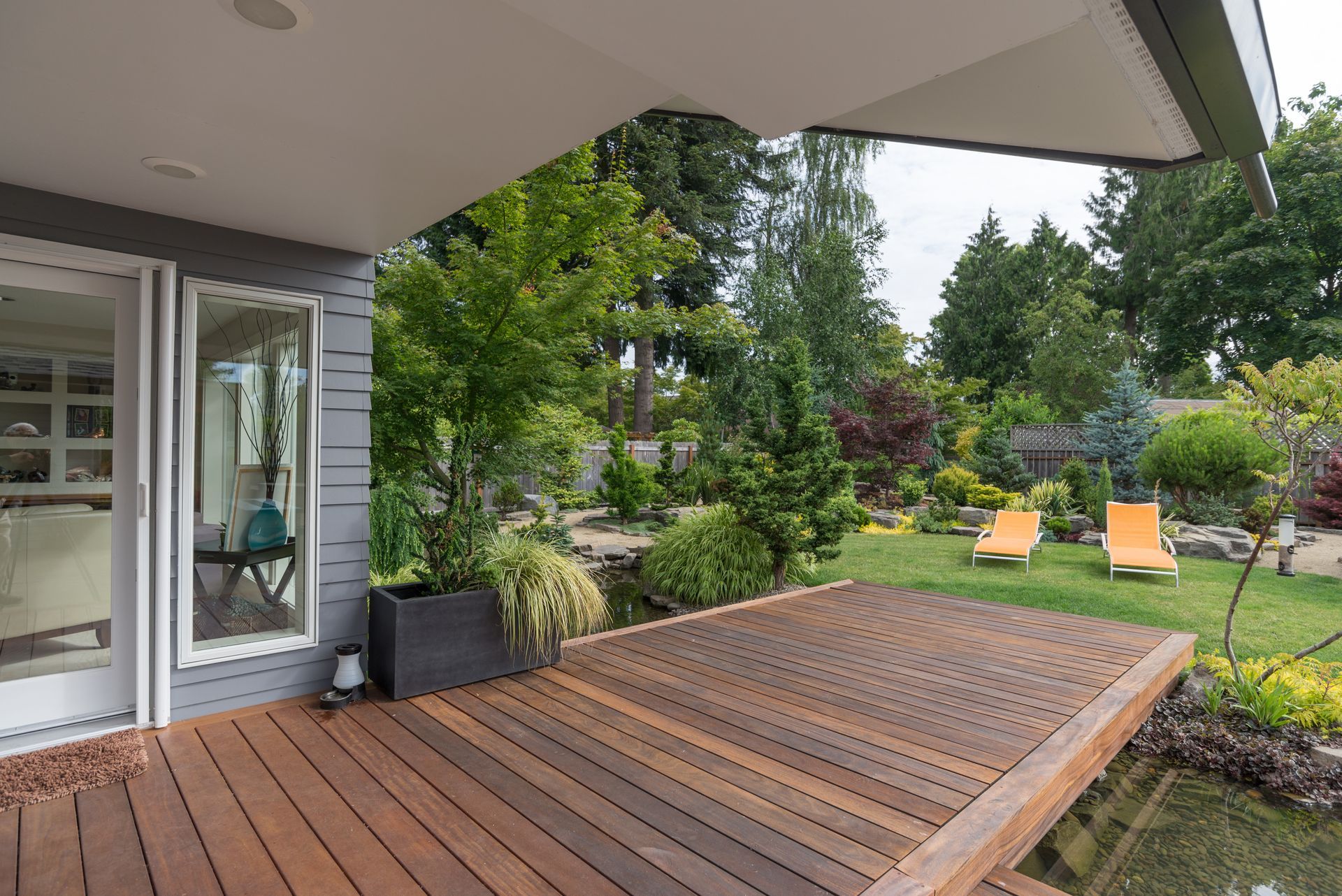 Wooden deck beside a house, overlooking a lush garden with two yellow chairs and trees.