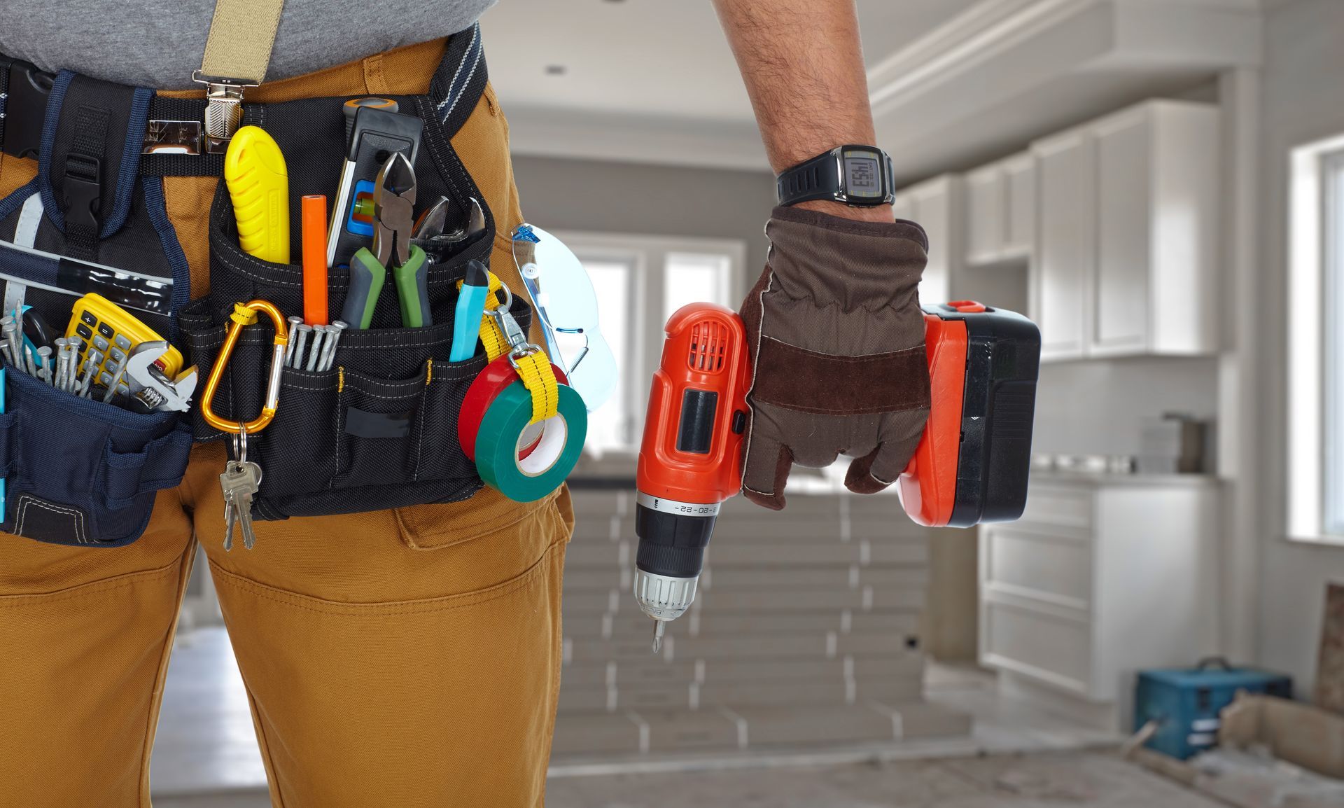 A man is holding a drill in a kitchen while wearing a tool belt.