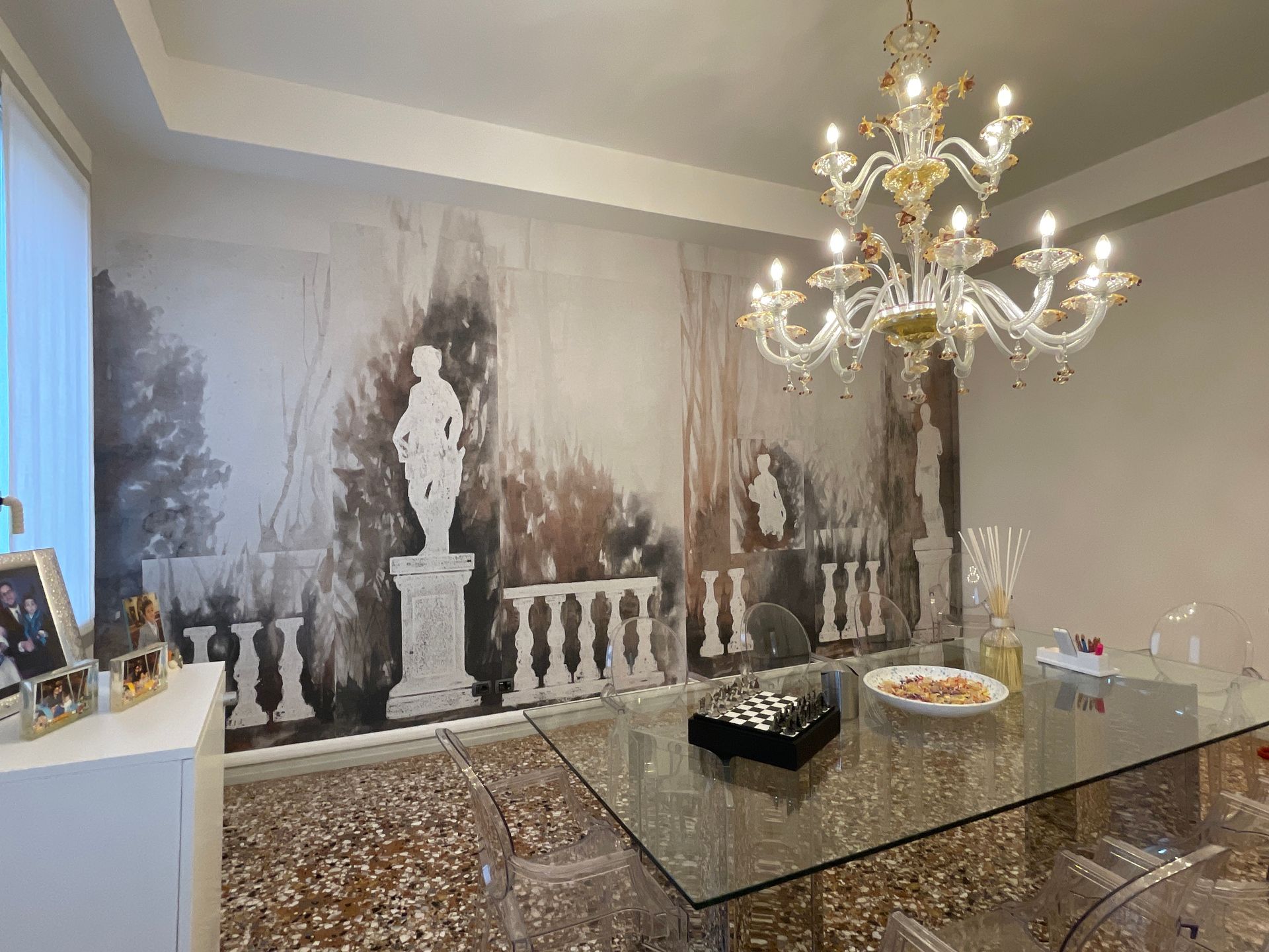 Sala da pranzo con lampadario, murale, tavolo in vetro e sedie trasparenti.