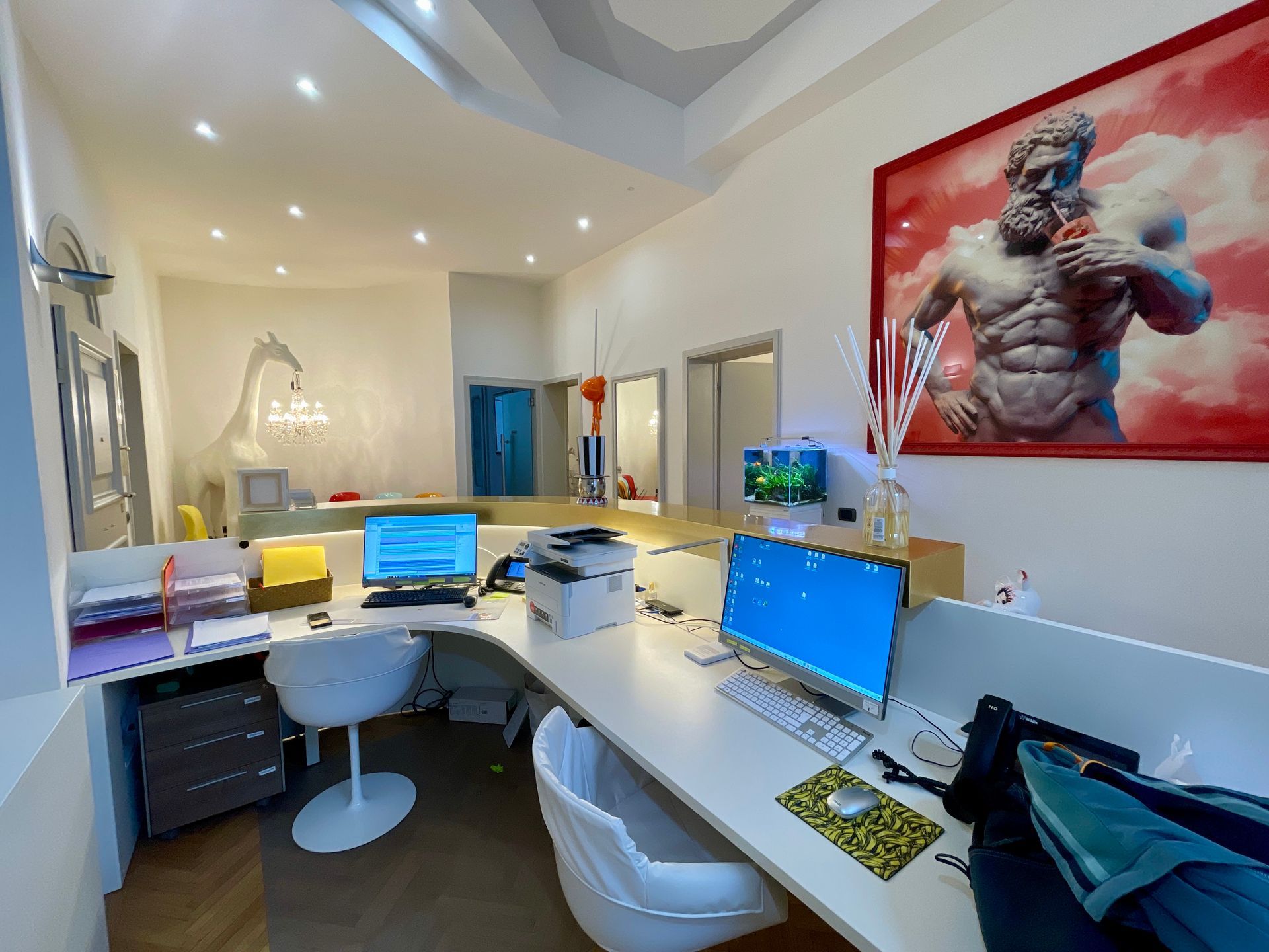 Uno spazio di lavoro con monitor per computer, stampante e un grande dipinto raffigurante una statua.