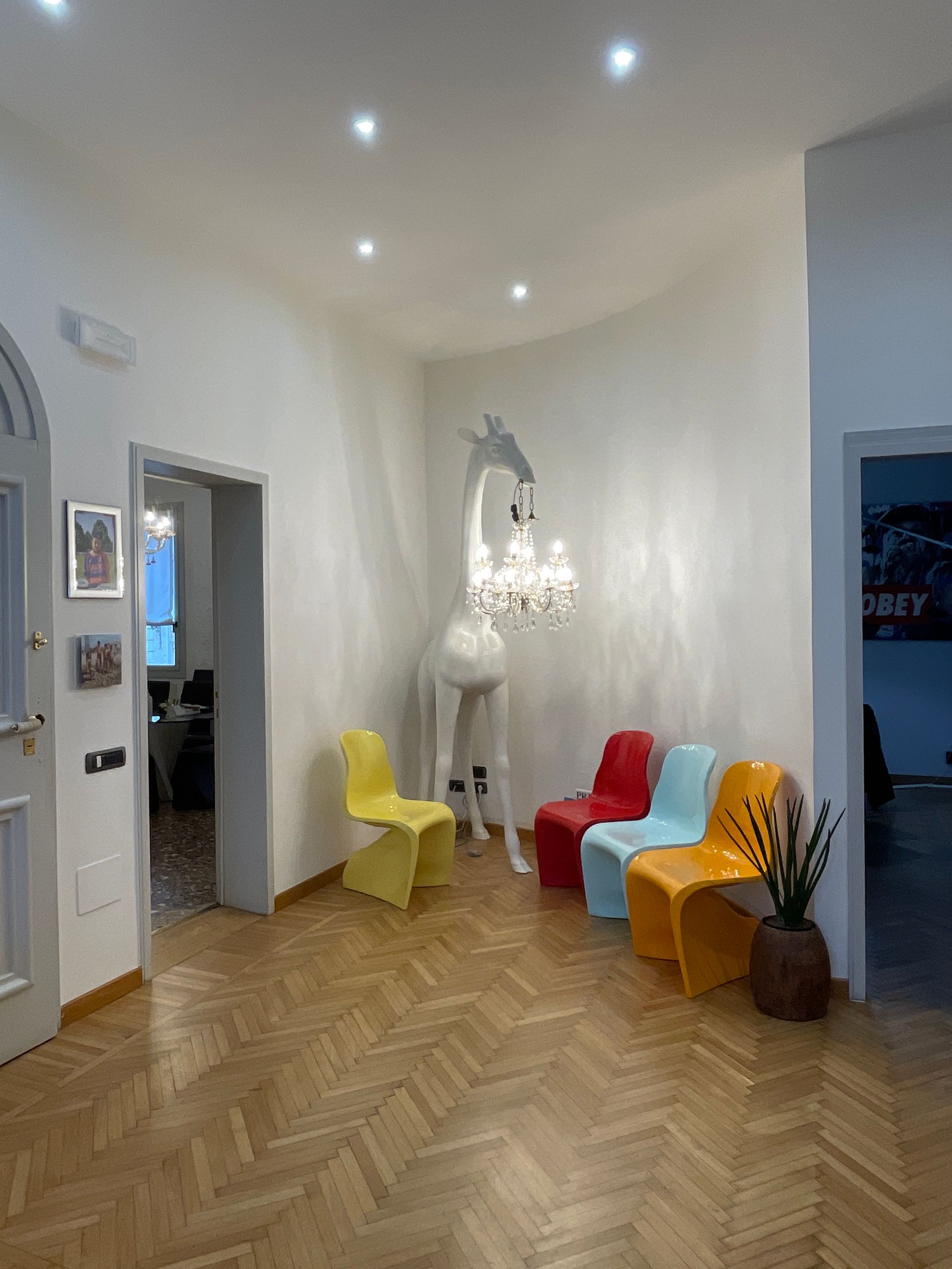 Sala d'attesa con lampadario a forma di giraffa e sedie colorate su pavimento a spina di pesce.