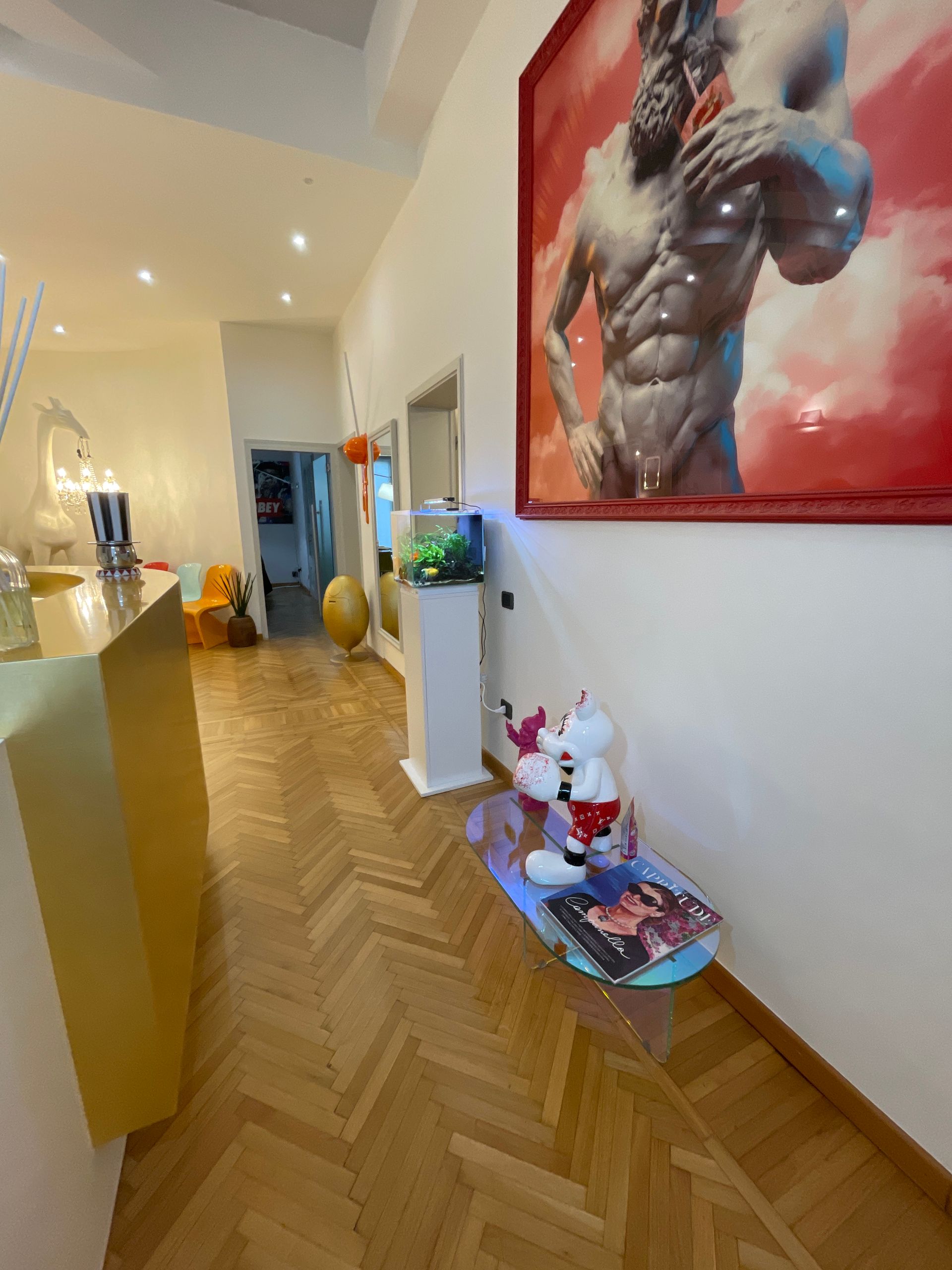 Corridoio con pavimento in legno, grandi quadri, piccolo acquario e una panca decorativa.