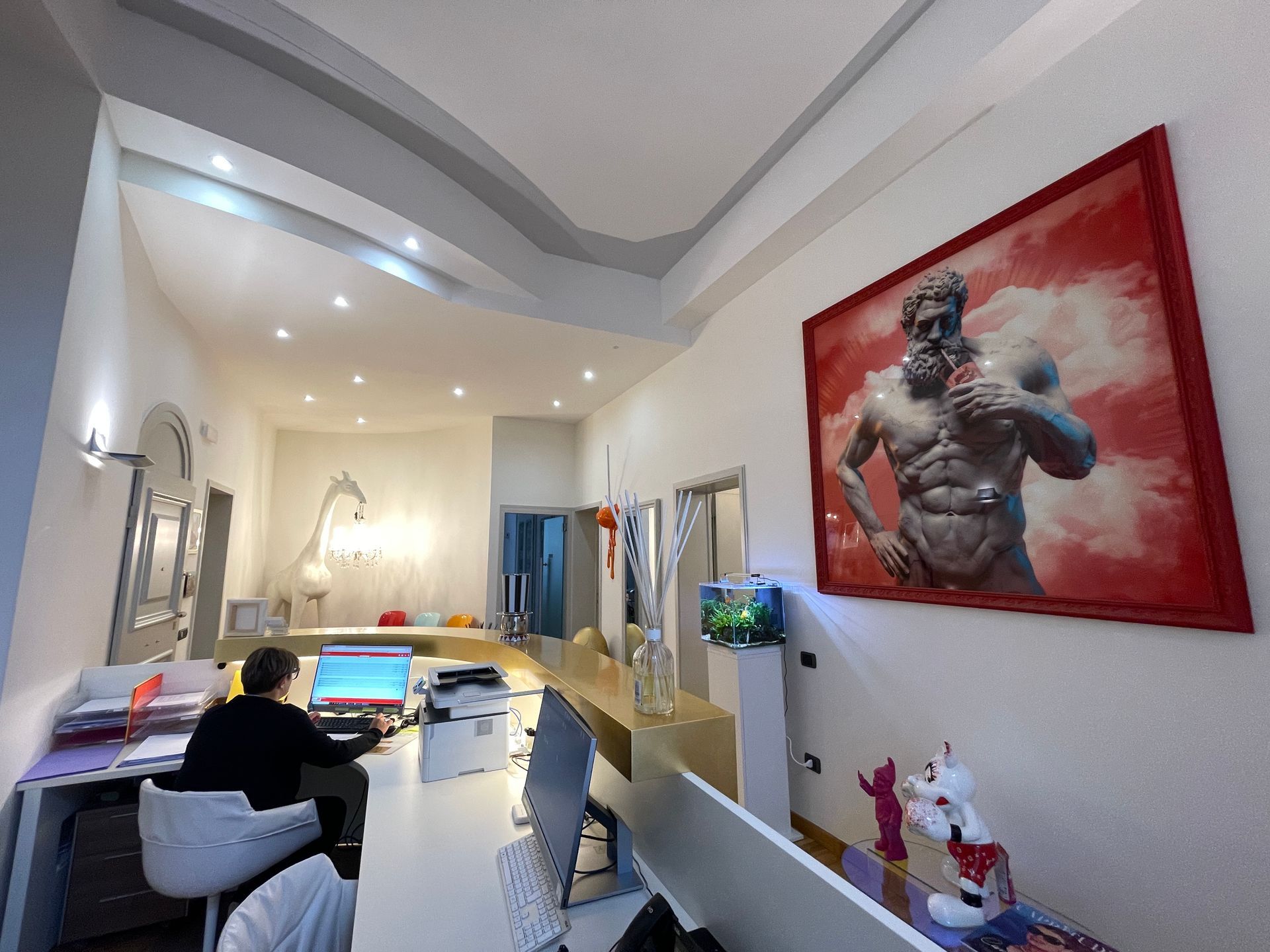 Interno di un ufficio con una persona al computer, quadri alle pareti e oggetti decorativi.