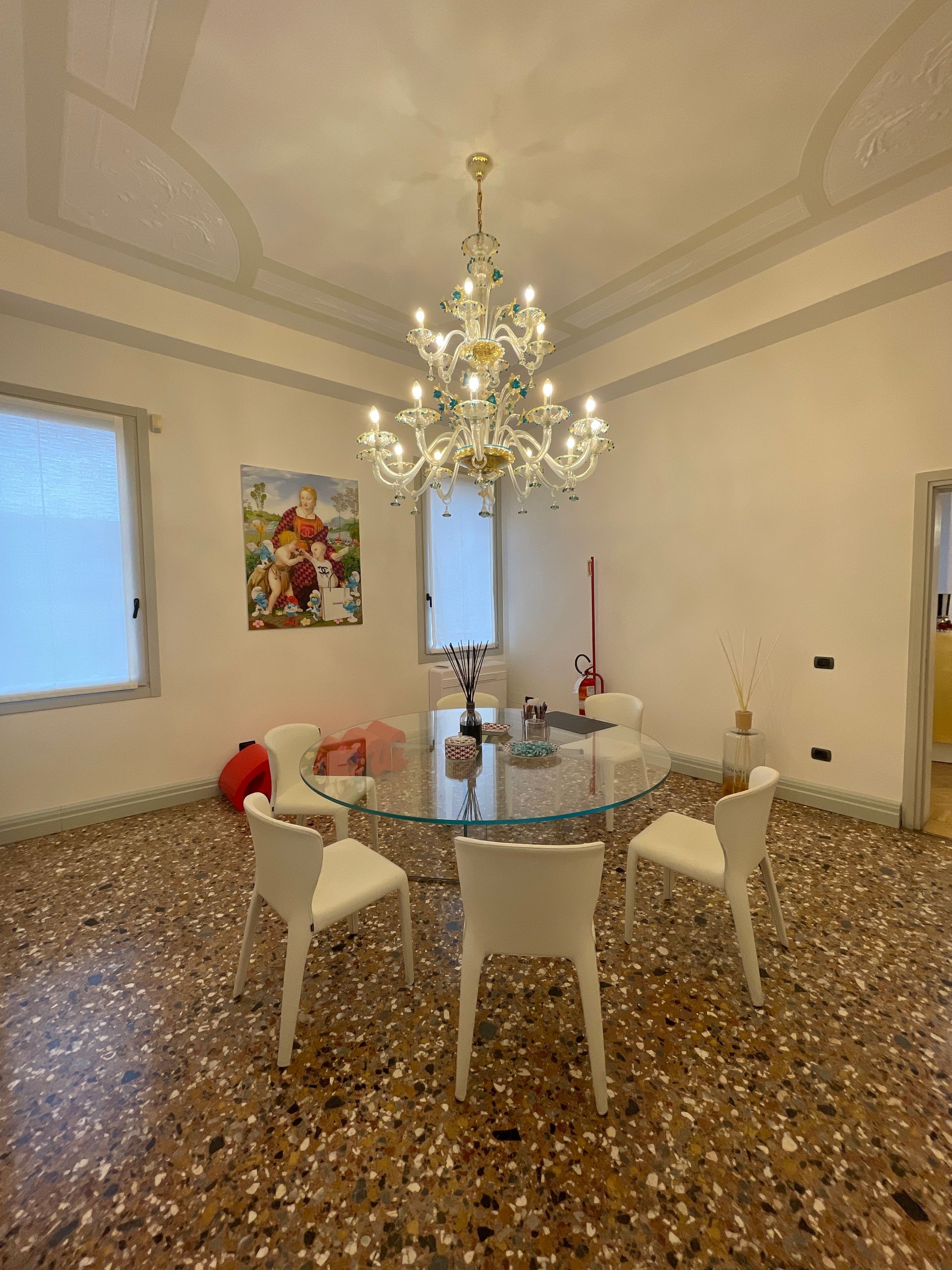 Tavolo rotondo in vetro con sedie bianche, lampadario, opere d'arte e pavimento maculato.