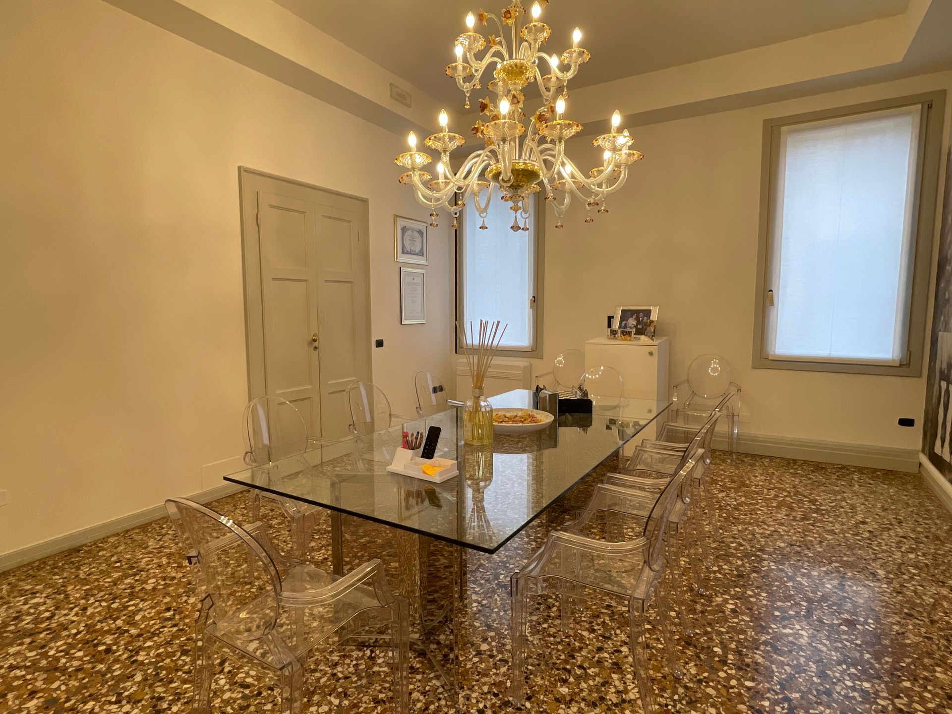 Sala da pranzo con tavolo in vetro, sedie trasparenti e lampadario decorato. Pavimento beige screziato e pareti chiare.