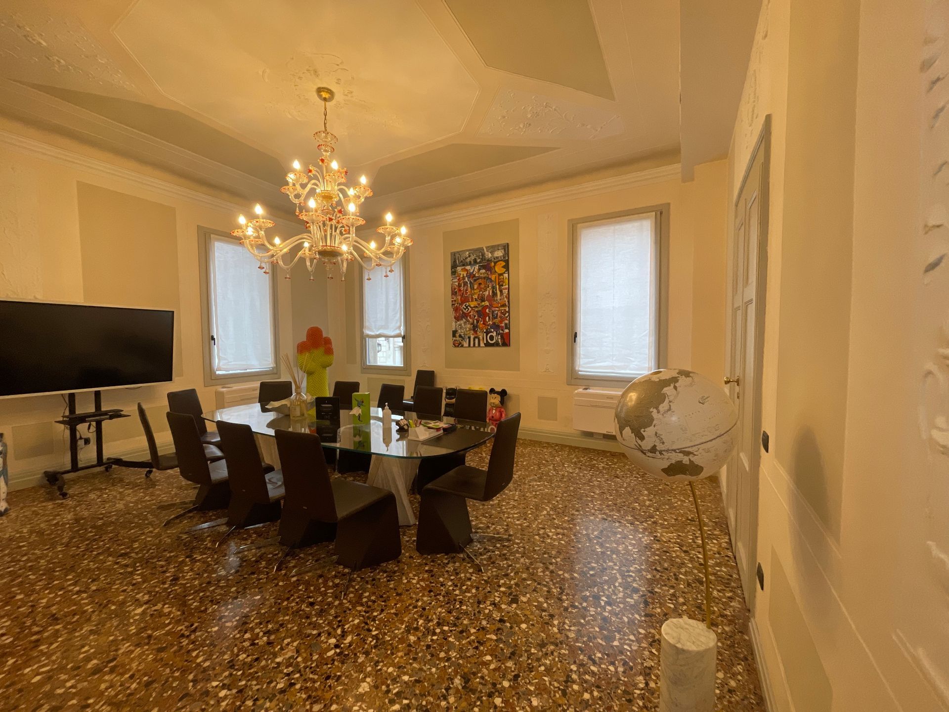 Sala conferenze con grande tavolo ovale, sedie, lampadario e opere d'arte. Finestre con tende. Pavimento in terrazzo.