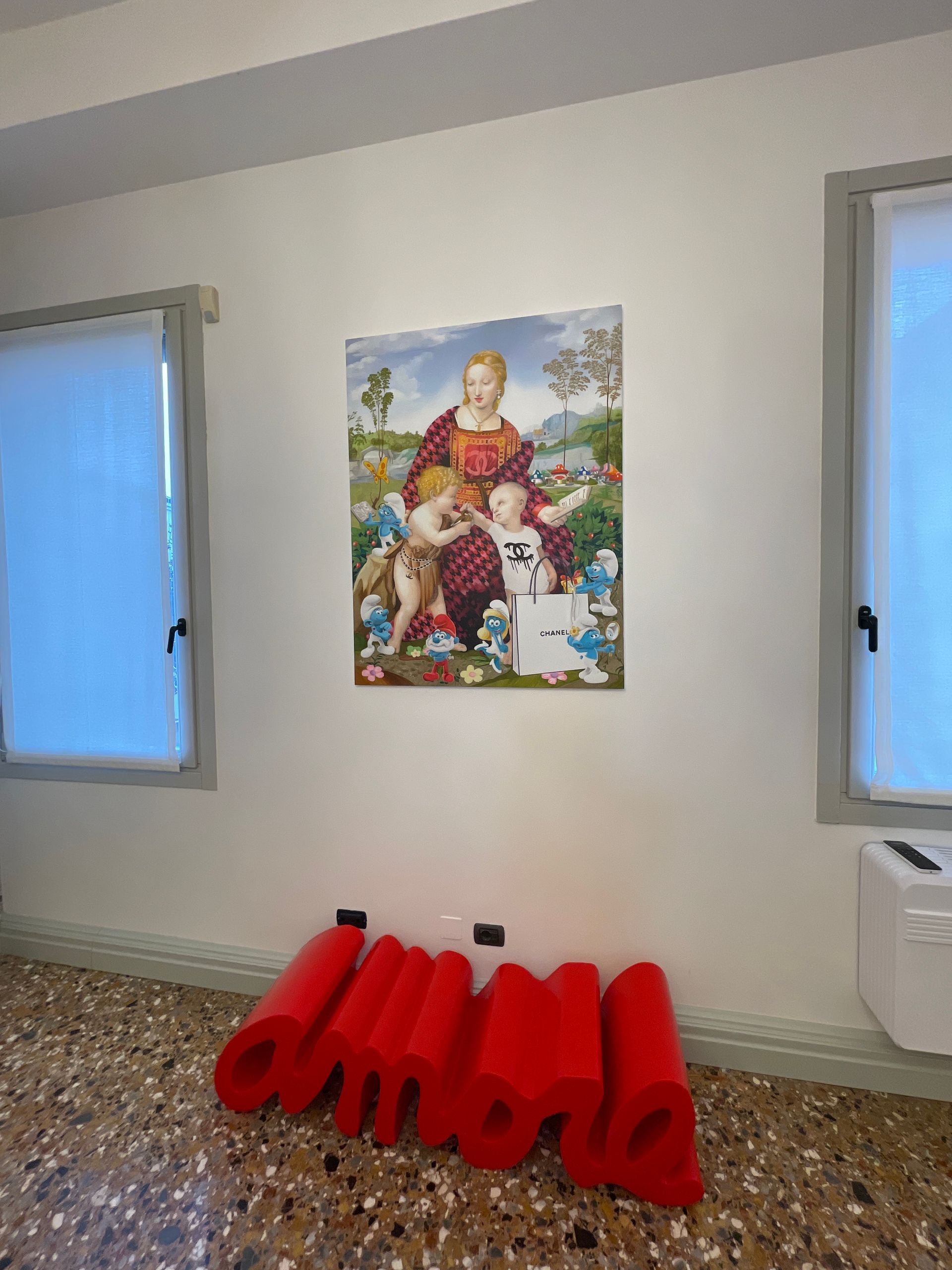 Opera d'arte raffigurante una donna e dei bambini circondati da figure blu, sopra un'installazione artistica rossa sul pavimento. Pareti bianche, finestre.