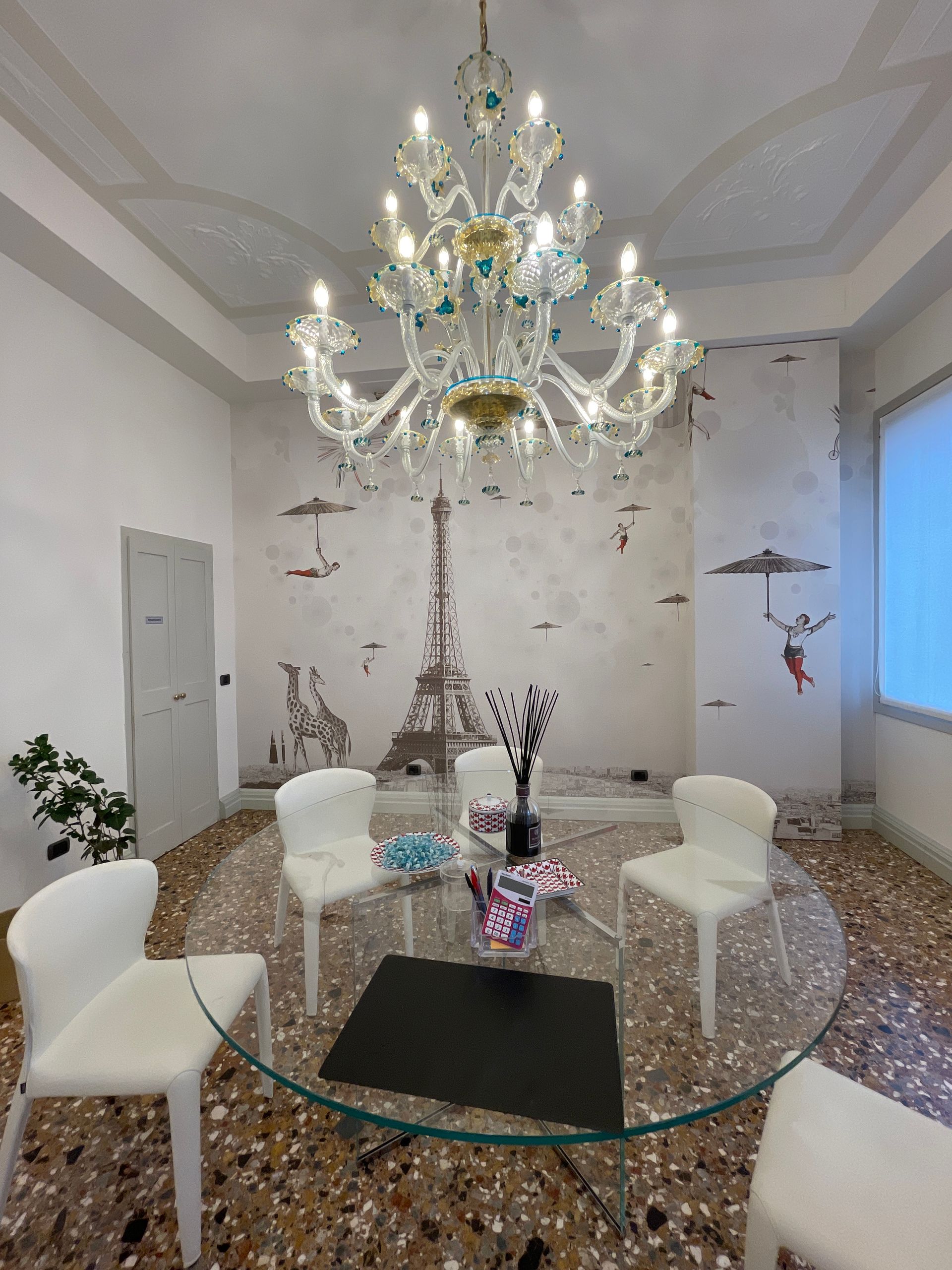 Una sala da pranzo con un tavolo in vetro, sedie bianche e un lampadario decorativo. Una parete è decorata con decorazioni in stile parigino.