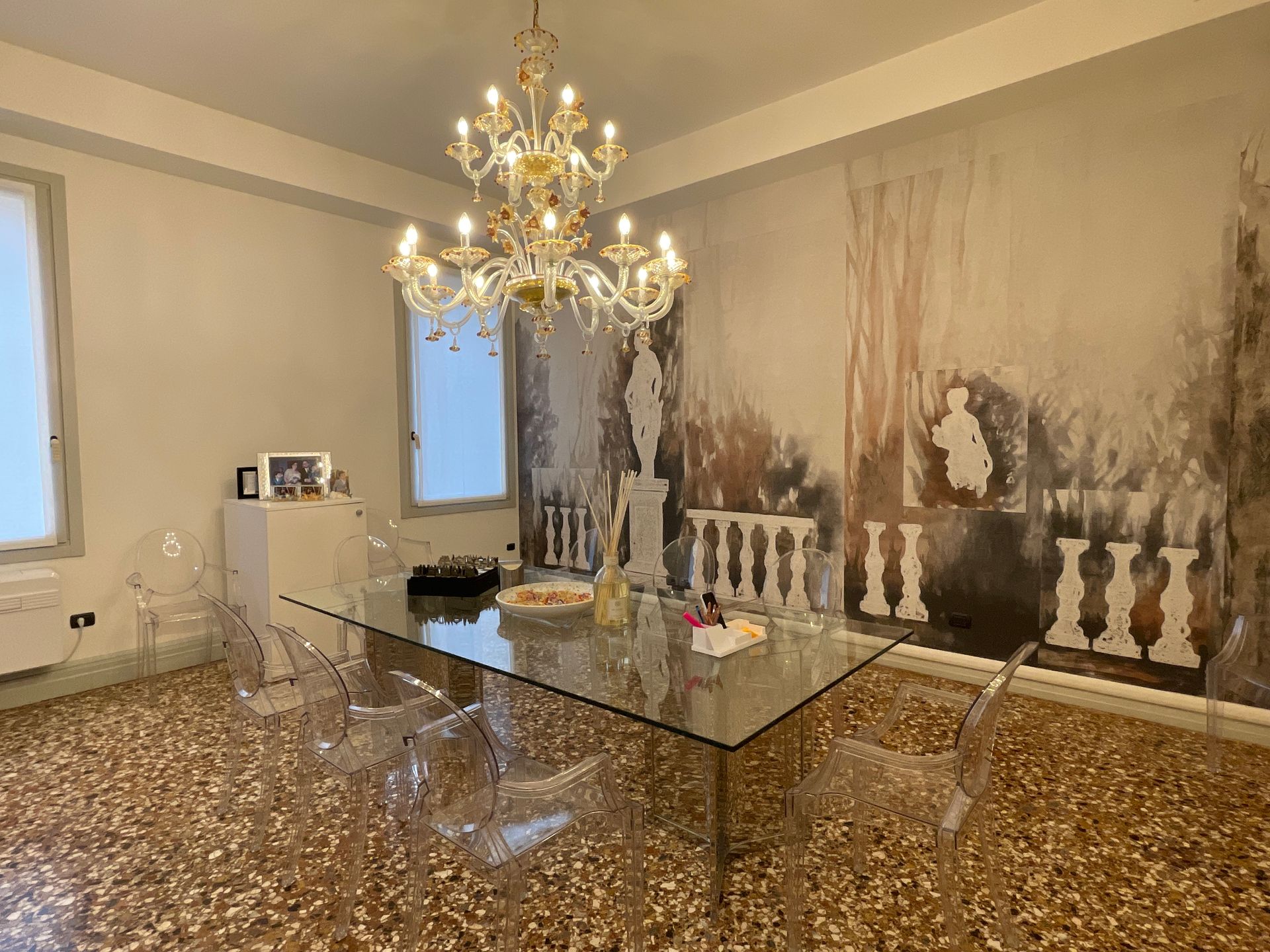 Sala da pranzo con tavolo in vetro, sedie trasparenti e un grande lampadario. Murale artistico sulla parete.