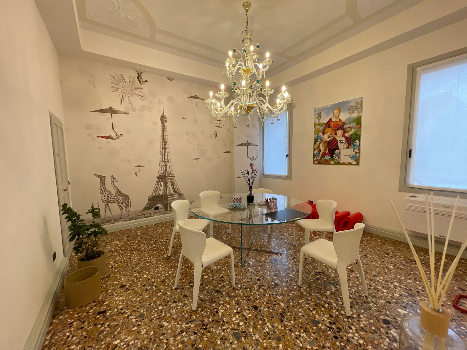 Sala da pranzo con tavolo in vetro, sedie bianche e lampadario. Un murale decora una parete.