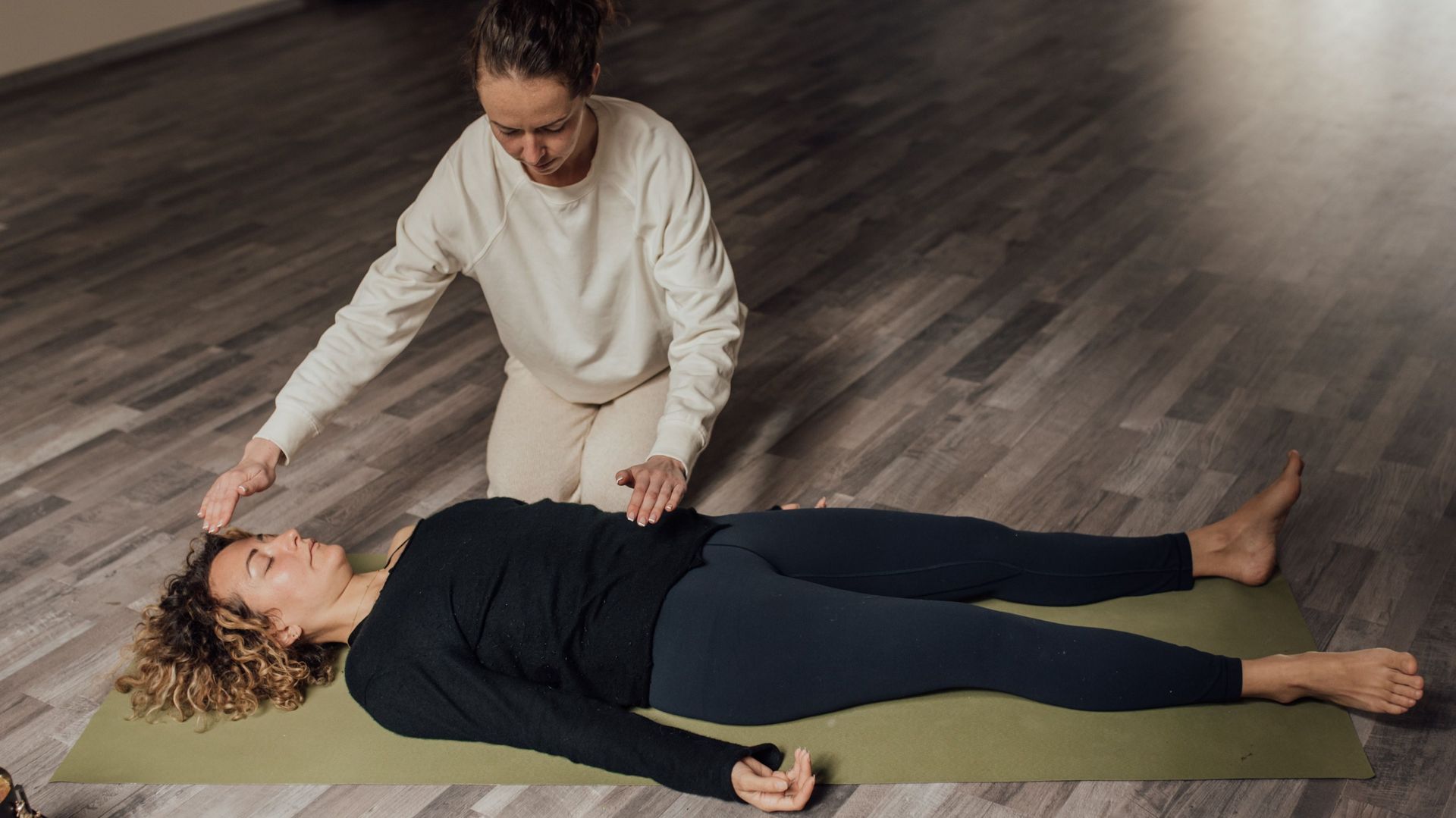 A woman is laying on a yoga mat while another woman looks on.