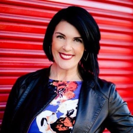 A woman wearing a leather jacket and a floral shirt is smiling in front of a red wall.