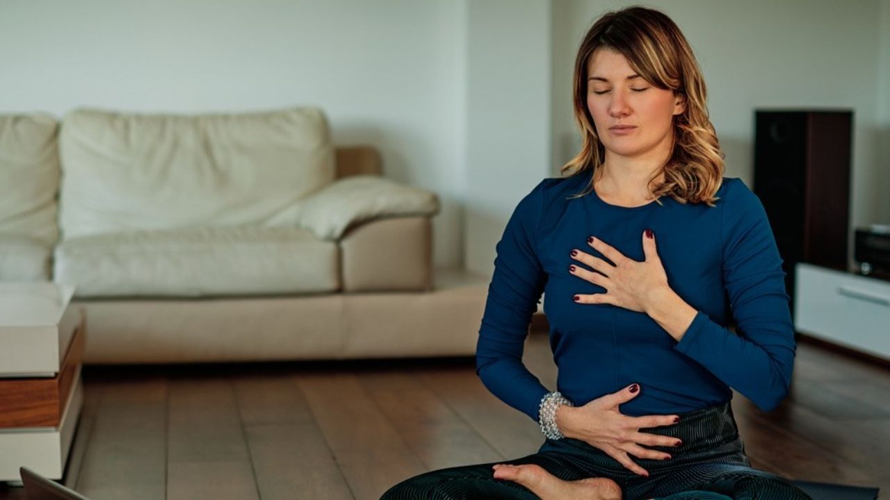 A woman is sitting in a lotus position on a yoga mat in a living room.