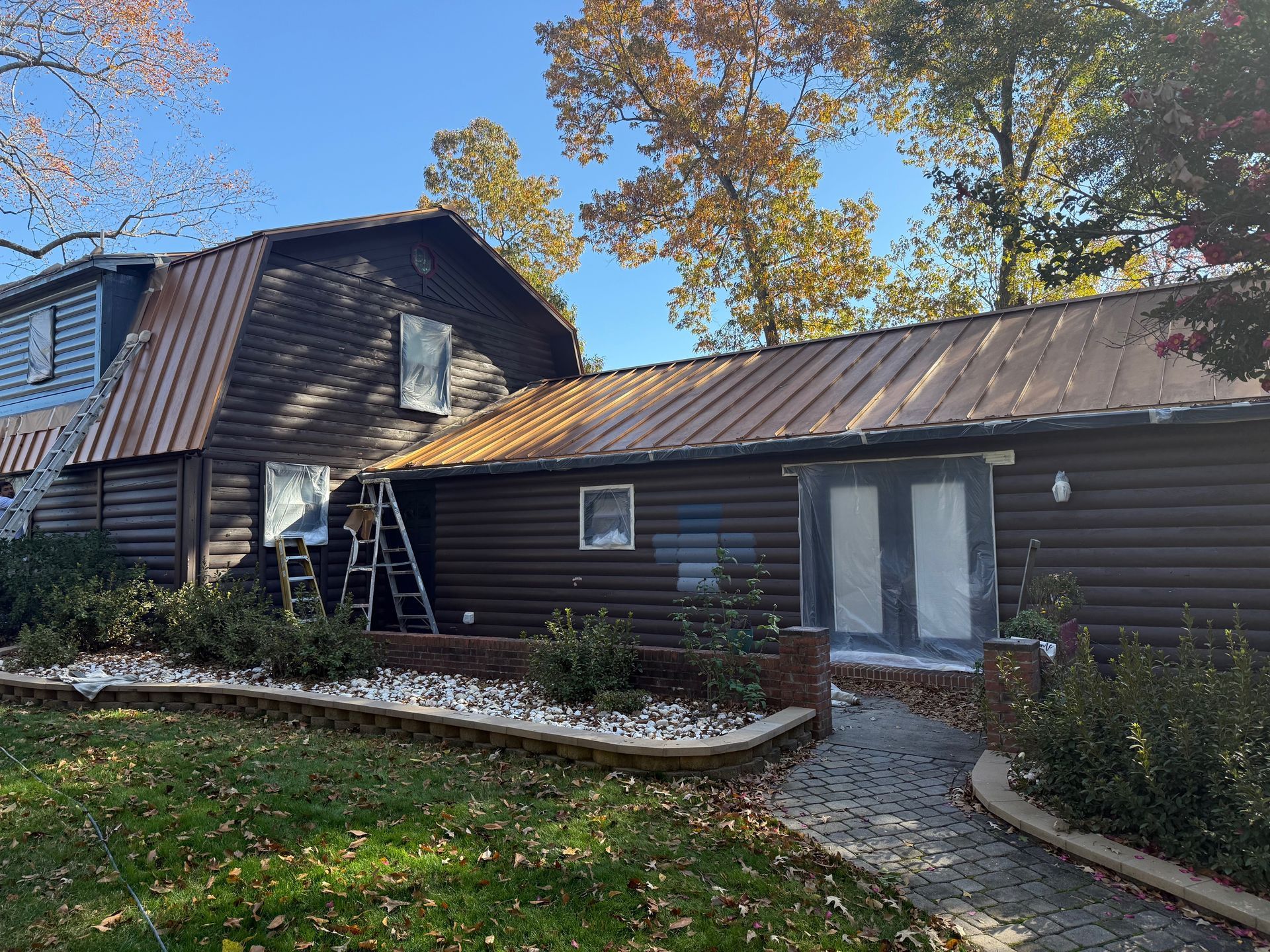 Brown log cabin being painted with a ladder visible. A person is on the ladder. Autumn trees and a blue sky.