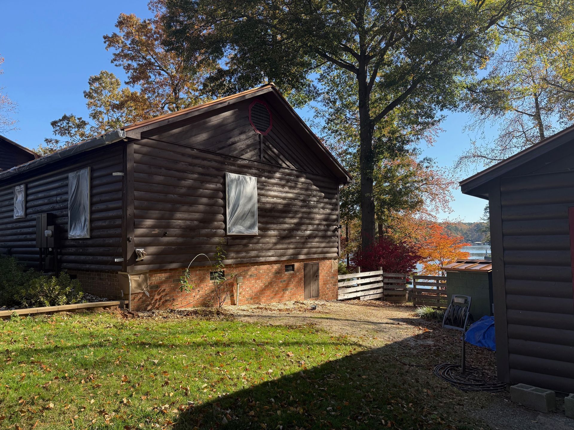Log cabins on a grassy lawn; a path leads towards a lake with fall foliage and a blue sky.