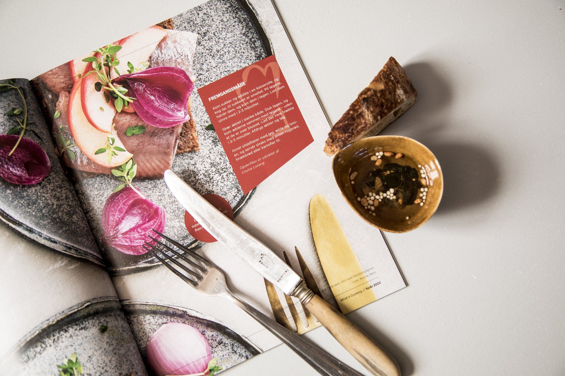 Et stemningsfuldt opslag i magasinet What's Cooking, hvor der er vist en lækker ret af en sildemad. På billeder er også et flot rustikt bestiksæt med en skive brød.