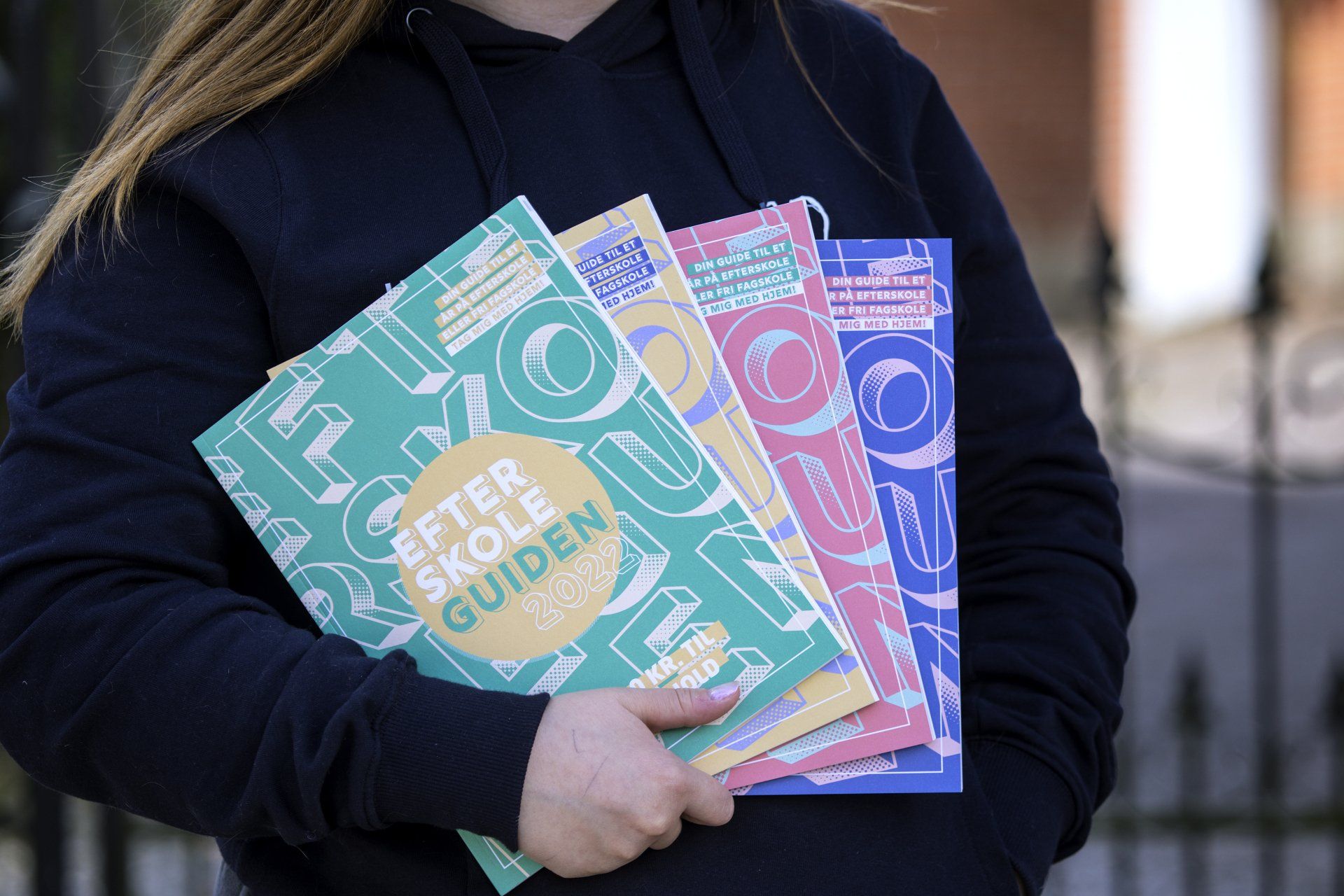 Billedet viser en kvinde, der holder fire udgaver af Efterskoleguiden i højre hånd. Hun holder magasinerne ind mod kroppen. Der er en grøn, gul, rød og blå udgave af magasinet.