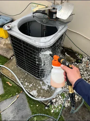 A person cleans an outdoor air conditioning unit's coils with a hose-attached foaming detergent sprayer.