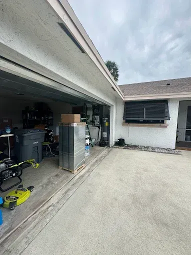 A wide view of a residential garage with a concrete driveway, metal shelving units, and a trash can inside.