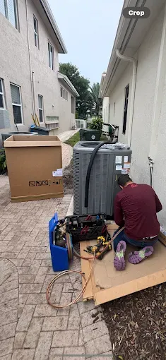 A technician in a maroon shirt kneels on a cardboard sheet while repairing an outdoor air conditioning unit.
