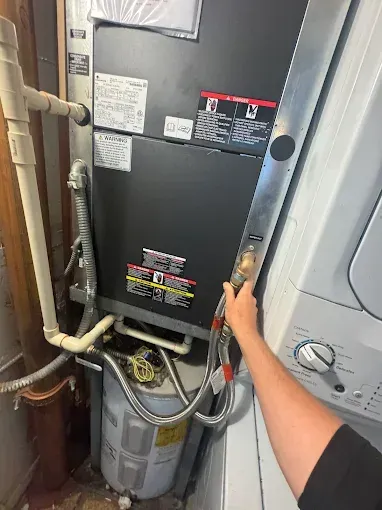 A person's hand reaches toward a metal hose connected to a black furnace unit next to a white laundry machine.