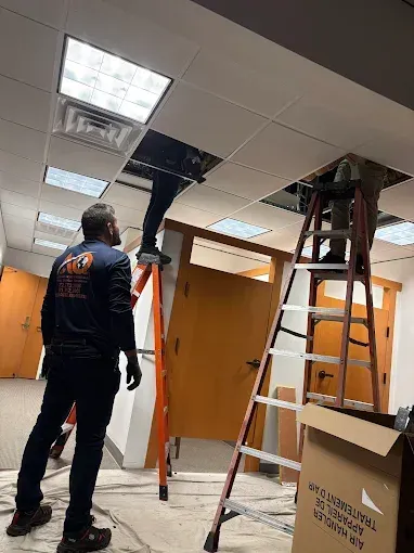 Two technicians on ladders work in an office ceiling plenum, while a third worker observes from the floor.