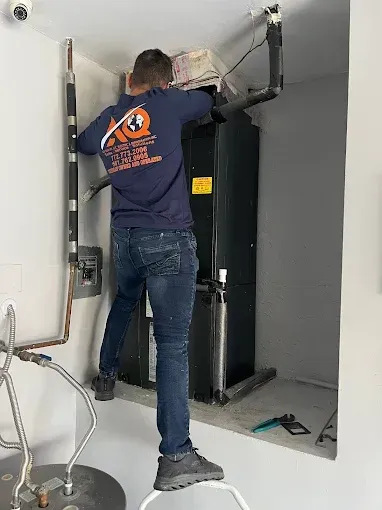 A technician in a blue uniform stands on a stool, working on HVAC equipment inside a wall alcove.