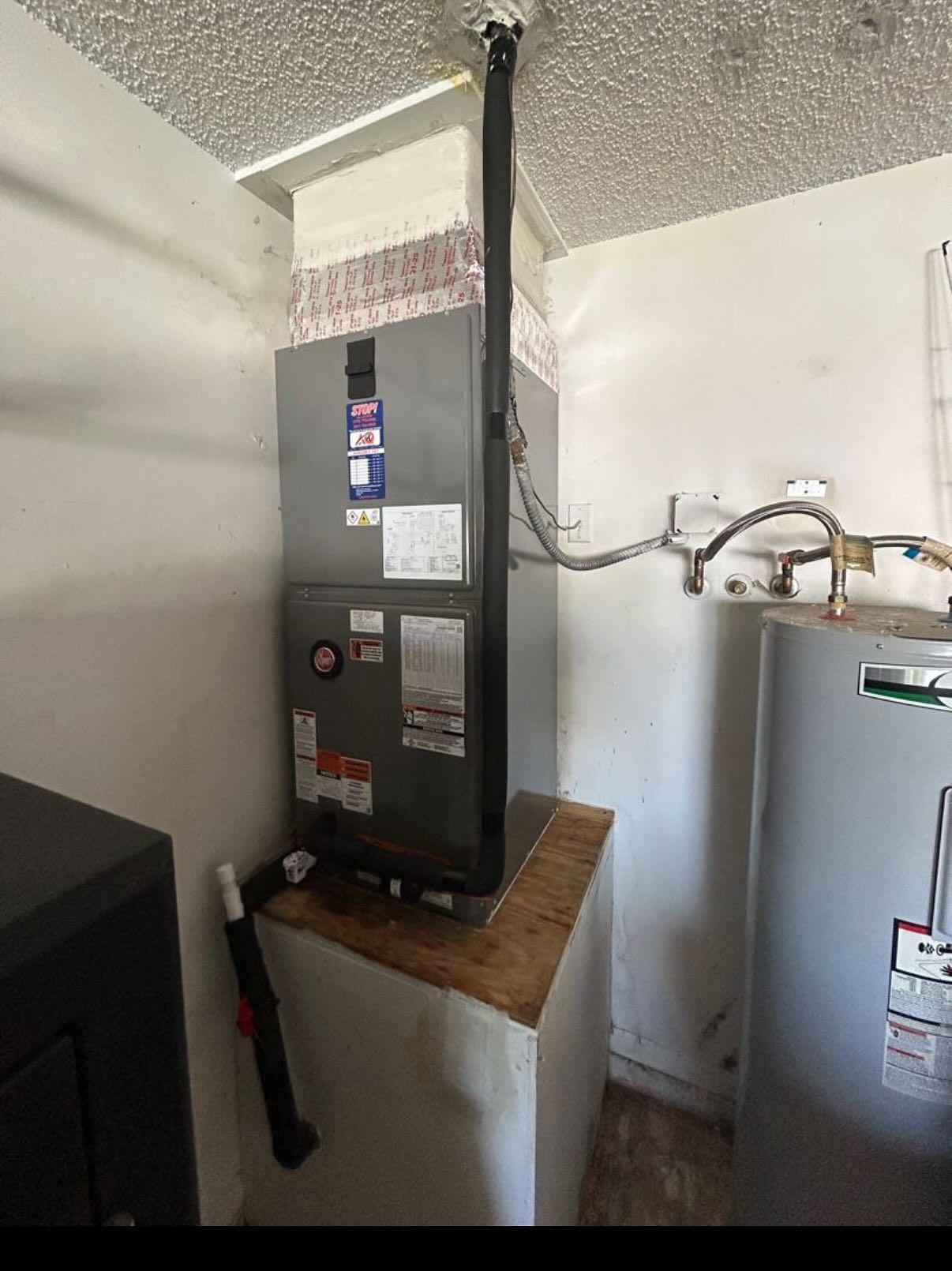 A vertical gas furnace sits on a white platform next to a water heater in a closet-like space.