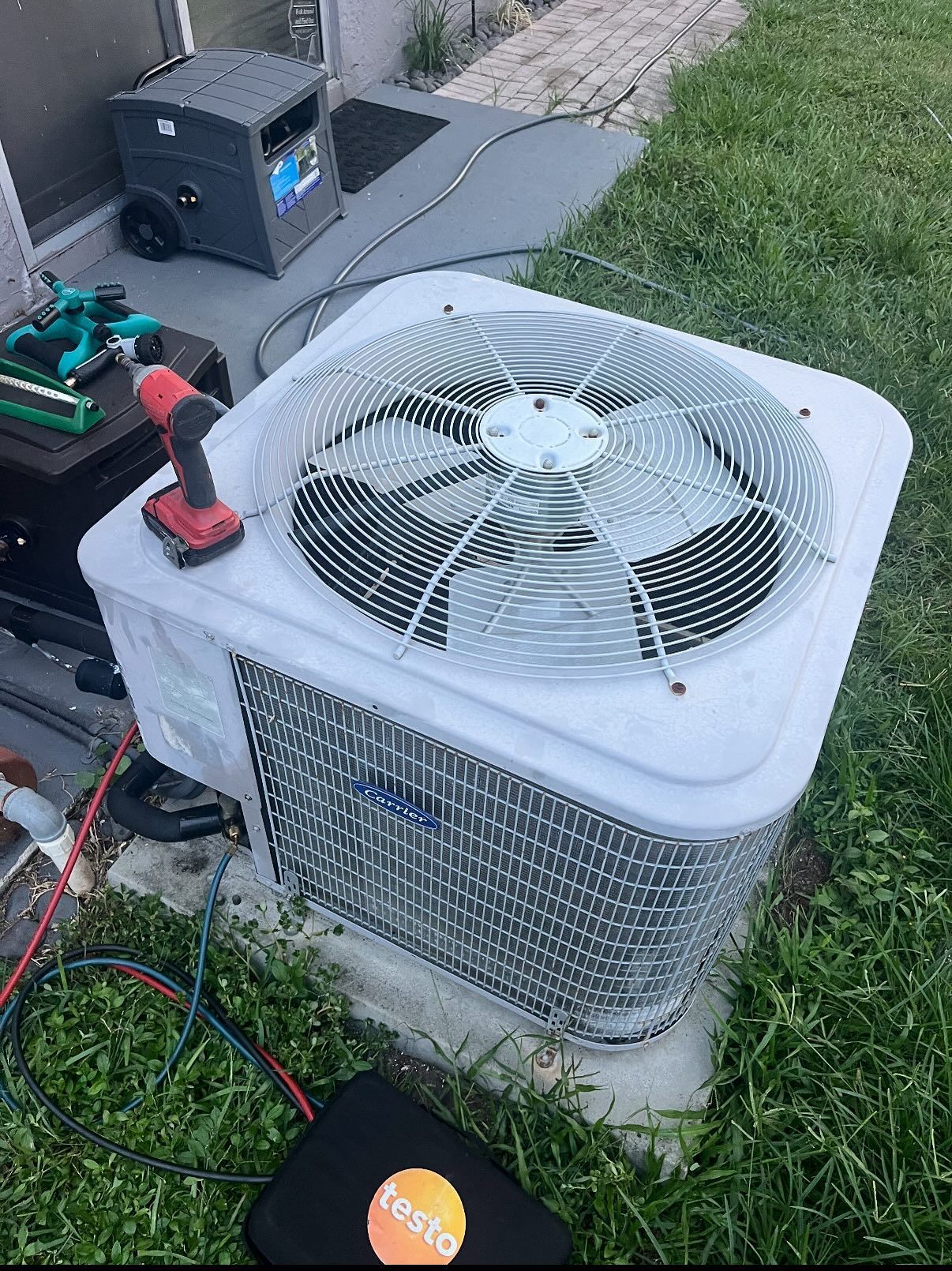 An outdoor HVAC unit being serviced, with diagnostic tools and a power drill on top, sitting on a concrete slab.
