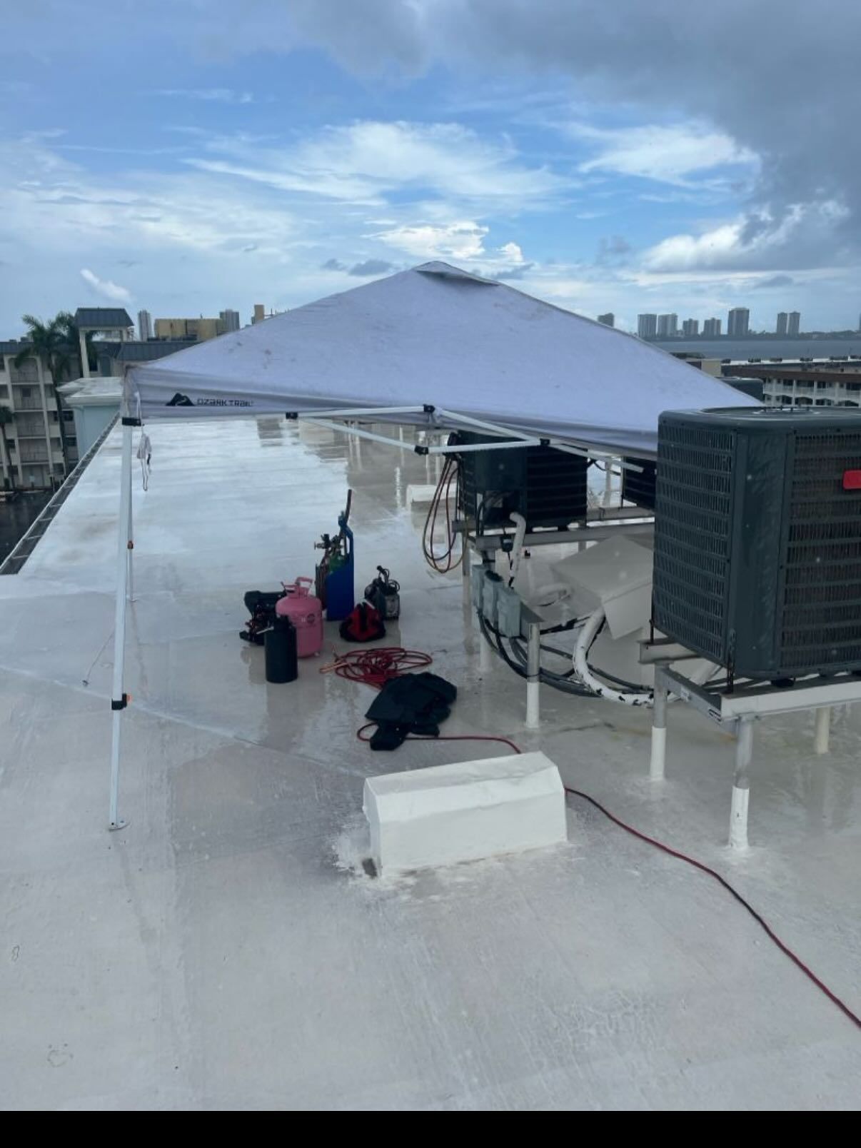A pop-up canopy shades tools and equipment, including a refrigerant tank, on a white rooftop near an AC unit.